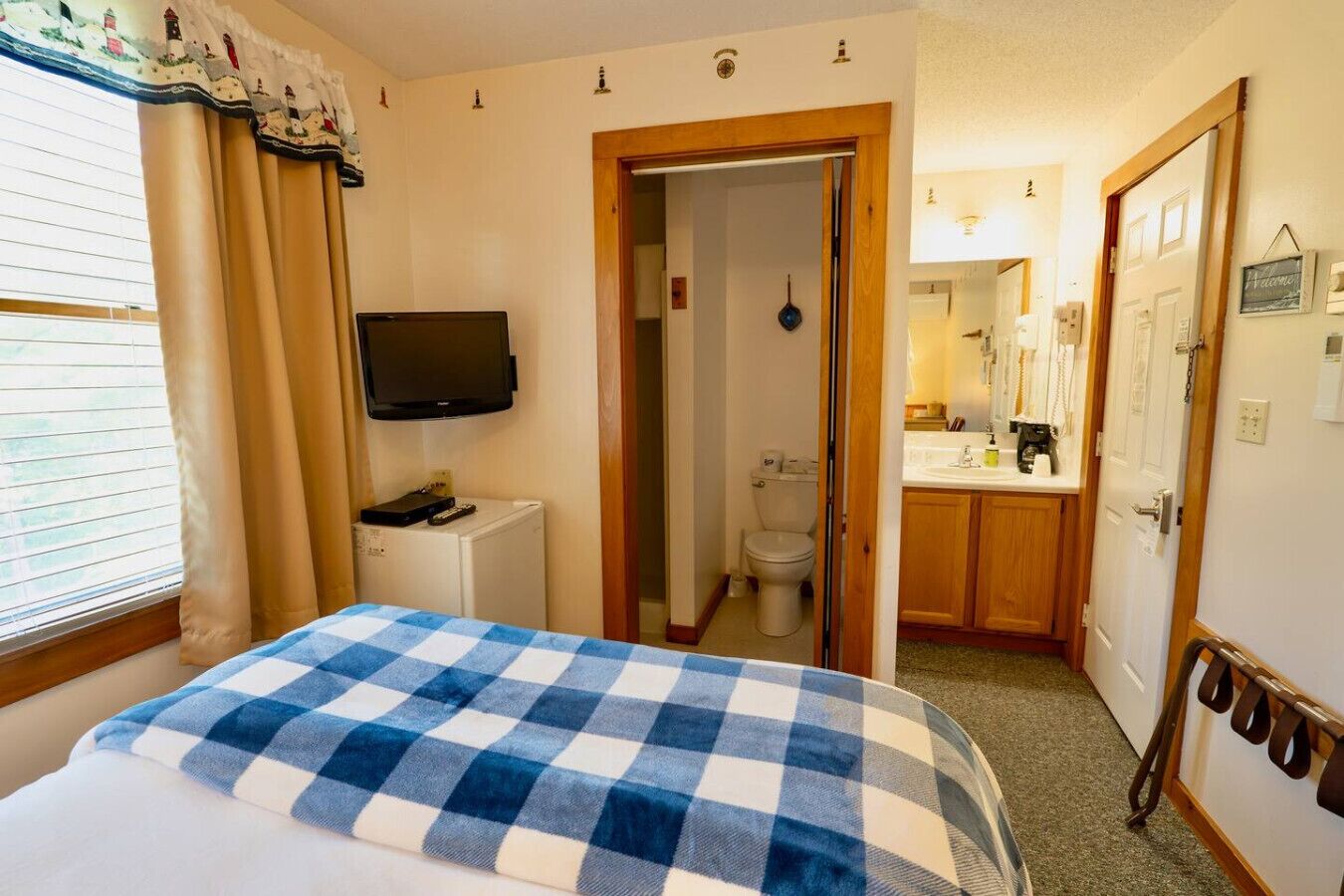 Hotellihuone, jossa on ruutukuviollinen peitto sängyllä, pienellä pöydällä oleva televisio, kylpyhuone, jossa on wc ja pesuallas, sekä puiset kaapit.