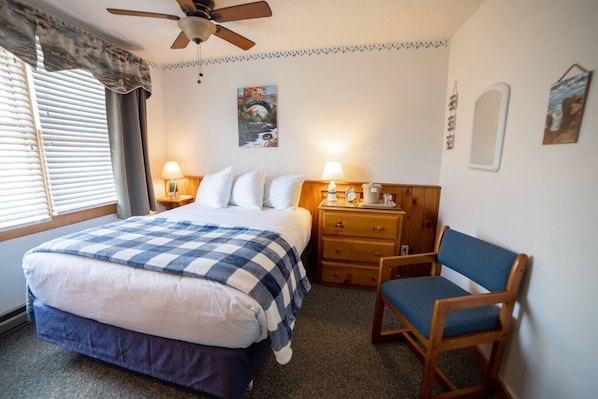 Relax in comfort with a large bed, nightstand, and chair, plus a ceiling fan and window with blinds.