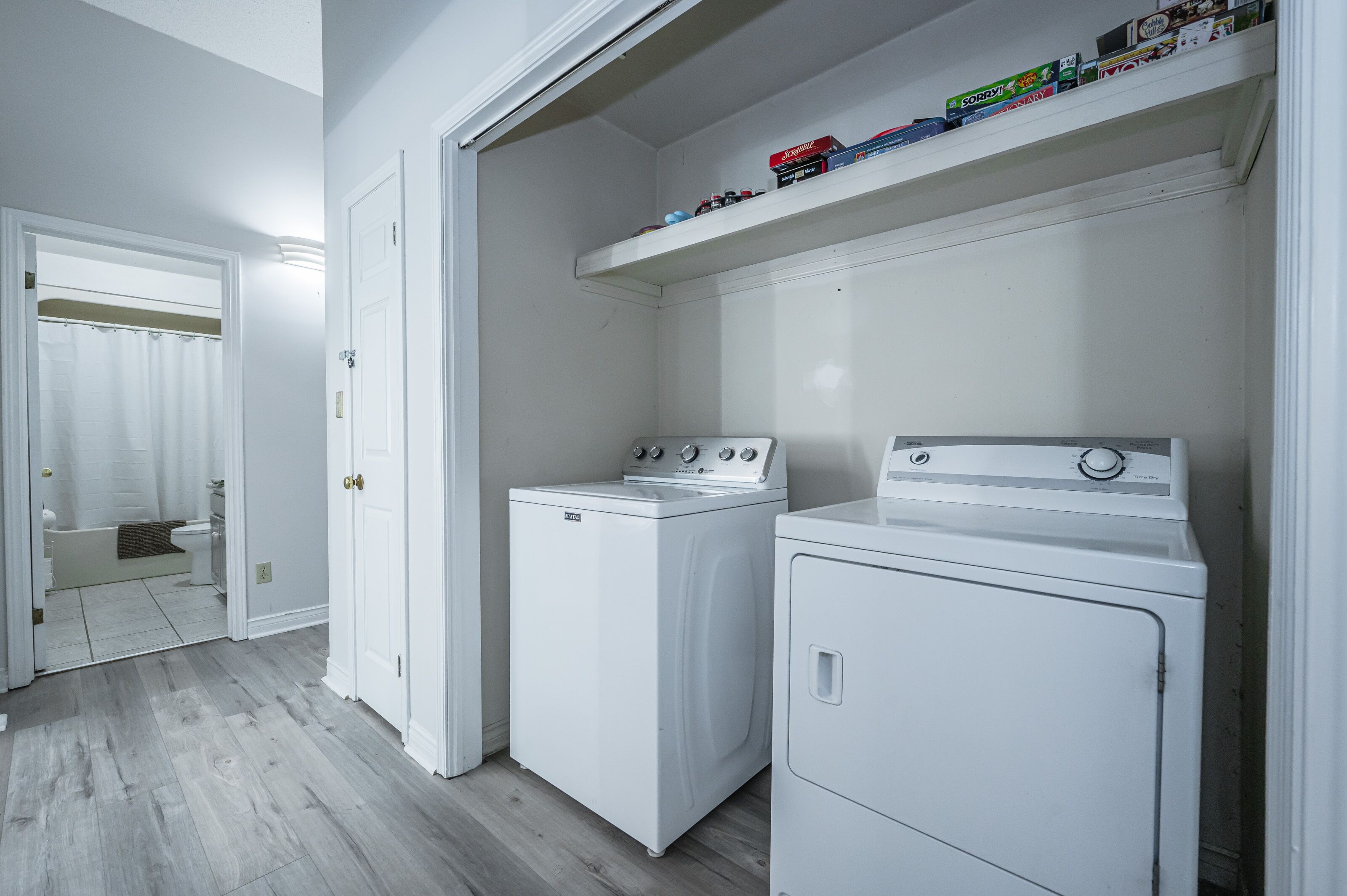 Une buanderie avec deux machines à laver et deux sèche-linge blancs, une porte blanche et une étagère au-dessus des sèche-linge.
