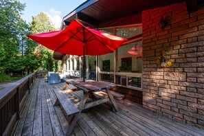 Una terraza de madera con una mesa de picnic y un paraguas rojo, con vistas a un edificio de ladrillo con ventanas amplias y un muro de ladrillo.