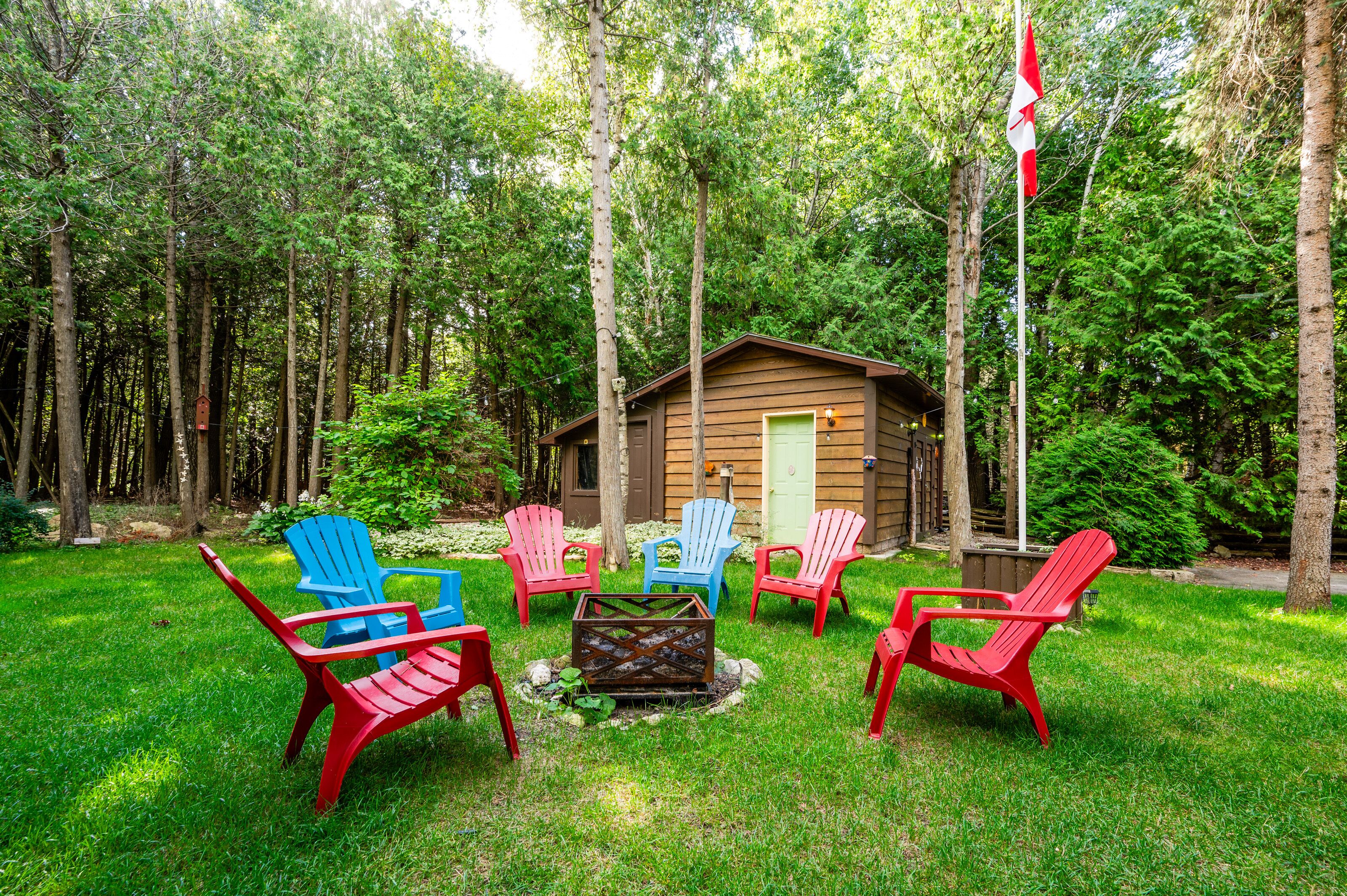 A cabin surrounded by a forest, with a fire pit and colorful Adirondack chairs in the yard.