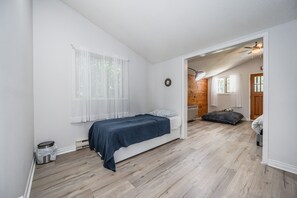 Un dormitorio con cama, una ventana con cortinas translúcidas, suelo de madera y una puerta que da a otra habitación.
