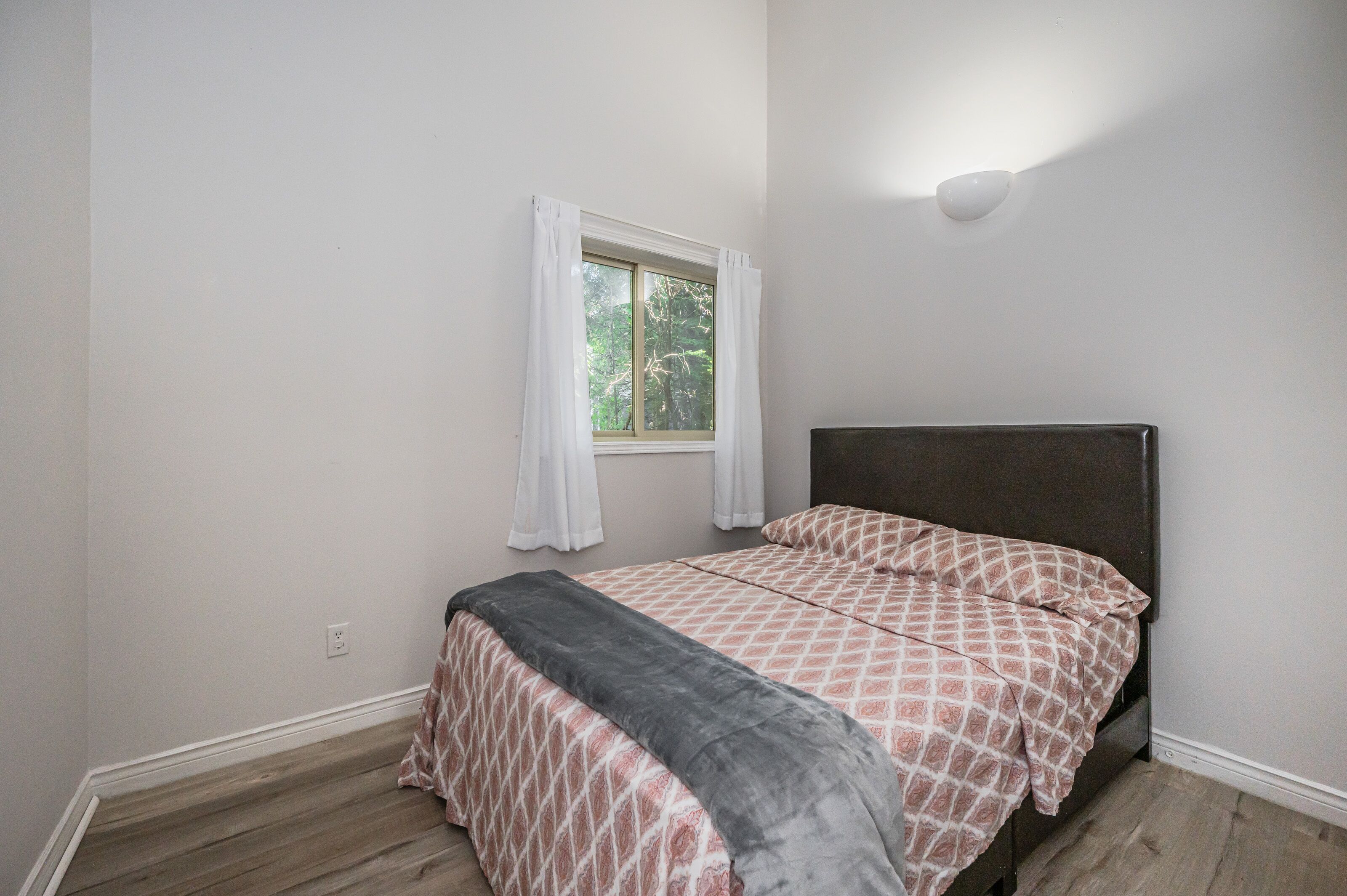 Une chambre à coucher avec un lit, une fenêtre avec des rideaux, un luminaire fixé au mur et un plancher en bois.
