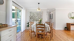 A dining area with a wooden table set for four, surrounded by wooden chairs. There is a large window with a view of greenery outside. - Bluebird Cottage – cheerful and cosy home, short distance to Bowral (Bowral)