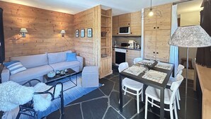 A cozy room with a sofa, dining table, chairs, a kitchen area, and a lamp. - Cozy studio near slopes - 4 guests (Méribel)