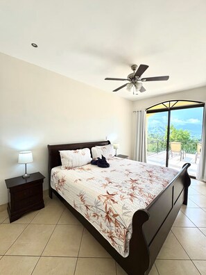 Une chambre à coucher comprenant un lit, des tables de chevet, un ventilateur de plafond et une vue sur un paysage montagneux par une grande fenêtre.