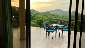 A room with a sliding glass door leading to an outdoor terrace with a table and chairs, a view of green hills, and a ceiling fan. - Seaside Serenity; 2b/2b with Panoramic Terrace (Playas Del Coco)