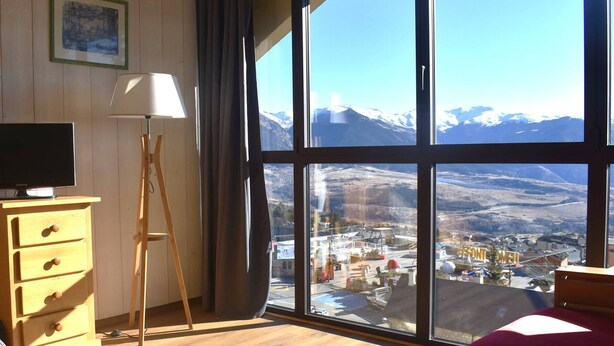A room with a large window offering a mountain view, a wooden dresser, a lamp, and a bed. - Charming 2-room cabin in central Font-Romeu, parking included (Font-Romeu-Odeillo-Via)