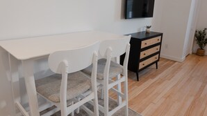 Een witte eettafel met twee stoelen, een zwarte dressoir met lades, een flatscreen televisie aan de muur en een potplant in de hoek.