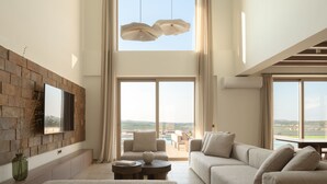 A spacious living room with a large flat-screen TV mounted on a stone wall, a comfortable sofa, a coffee table, and a view of a pool and landscape through large windows. - Casa Beleza Villa, a Mythical Retreat (Perama)