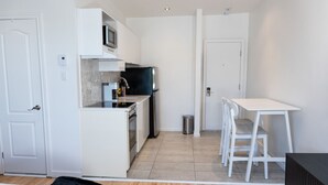 A compact kitchen with white cabinetry, a microwave, and a stove. There is a small trash bin and a white table with chairs. - #11 - Studio/Plateau/St-Denis/SelfCheck-In/AC/TV (Montréal)