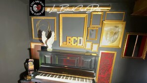 A piano with a black and gold finish, surrounded by framed artwork and decorative elements on the wall. - Stay & Play at Lovebehindcloseddoors (Plymouth)