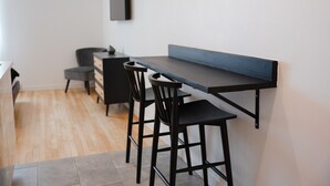 A modern kitchen with a dark countertop, two bar stools, a small cabinet, and a flat-screen TV mounted on the wall.