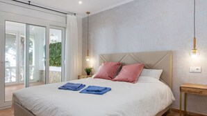 Une chambre dotée d’un grand lit, d’une table de chevet, d’une lampe fixée au mur et d’une porte-fenêtre coulissante donnant sur un espace extérieur.