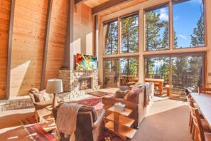 A cozy living room with a stone fireplace, large windows, and a view of the surrounding forest.