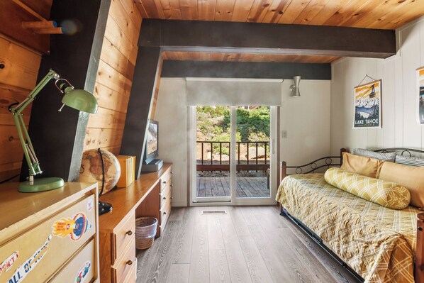 A bedroom with a wooden ceiling, a bed with patterned bedding, a desk with a lamp, and a sliding glass door leading to an outdoor deck.