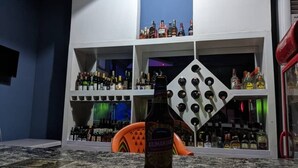 Una zona de bar con estantes llenos de botellas de licor, una botella de cerveza sobre la barra y un letrero de neón al fondo.