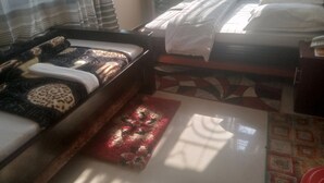 Una habitación con dos camas, una con cubrecama estampado y la otra con cubrecama blanco liso. Hay una ventana con cortinas, una mesita y un tapete estampado en el suelo.