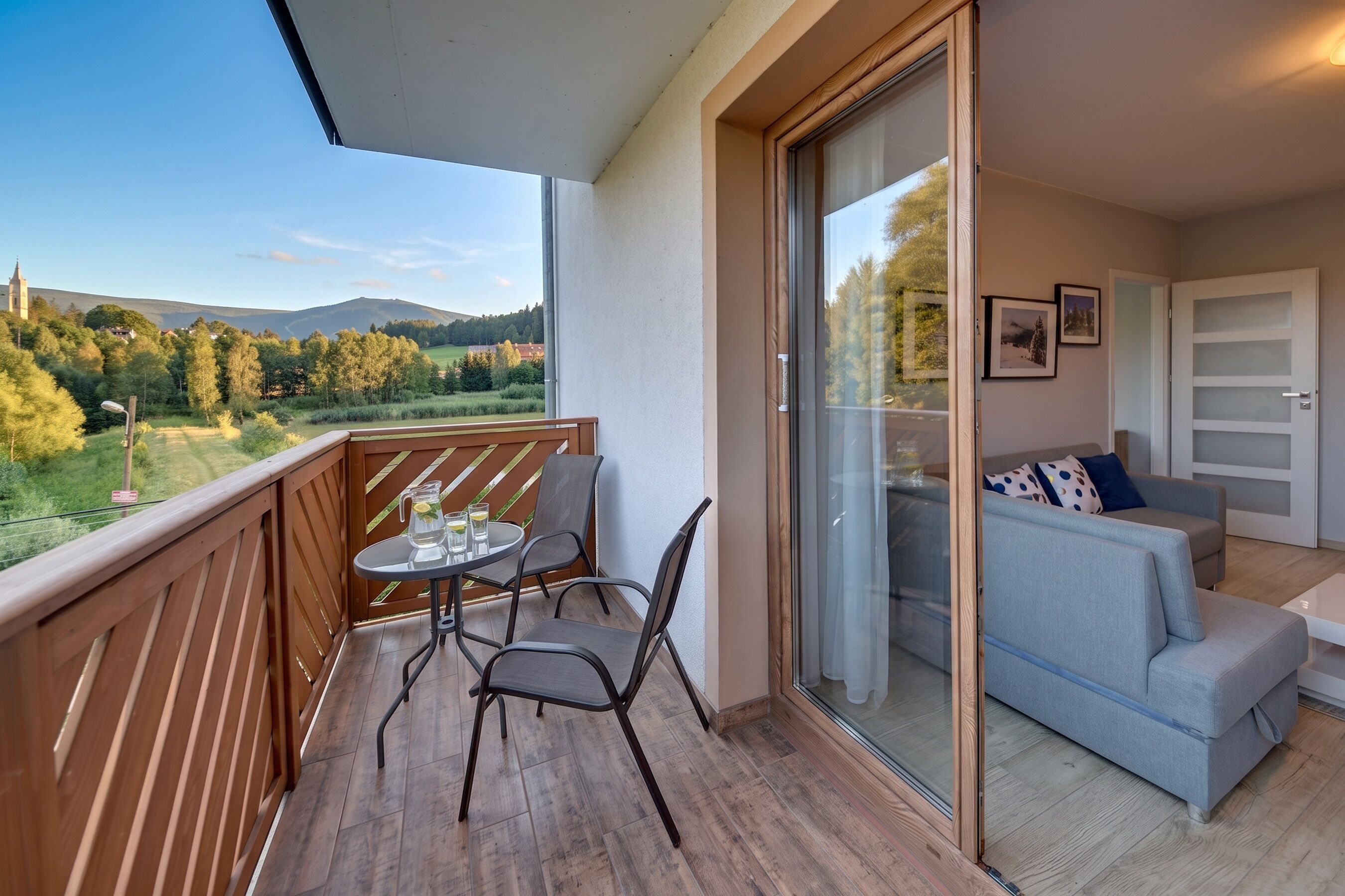 A balcony with a table and chairs, a sliding glass door leading to a living area with a sofa and cushions.