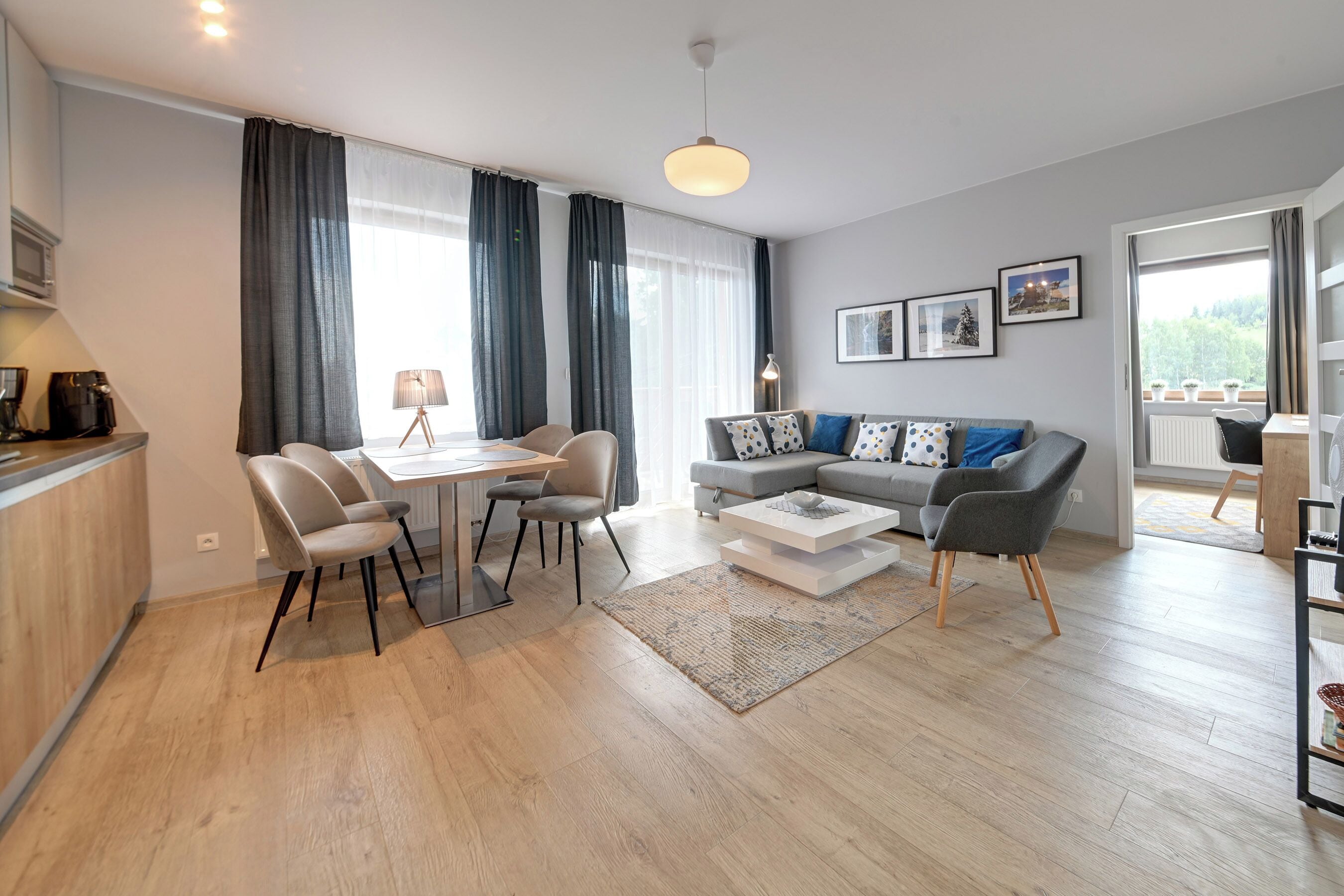 En moderne stue med spiseplads, en grå sofa med mønstrede puder, en kaffemaskine og et stort vindue med gardiner.