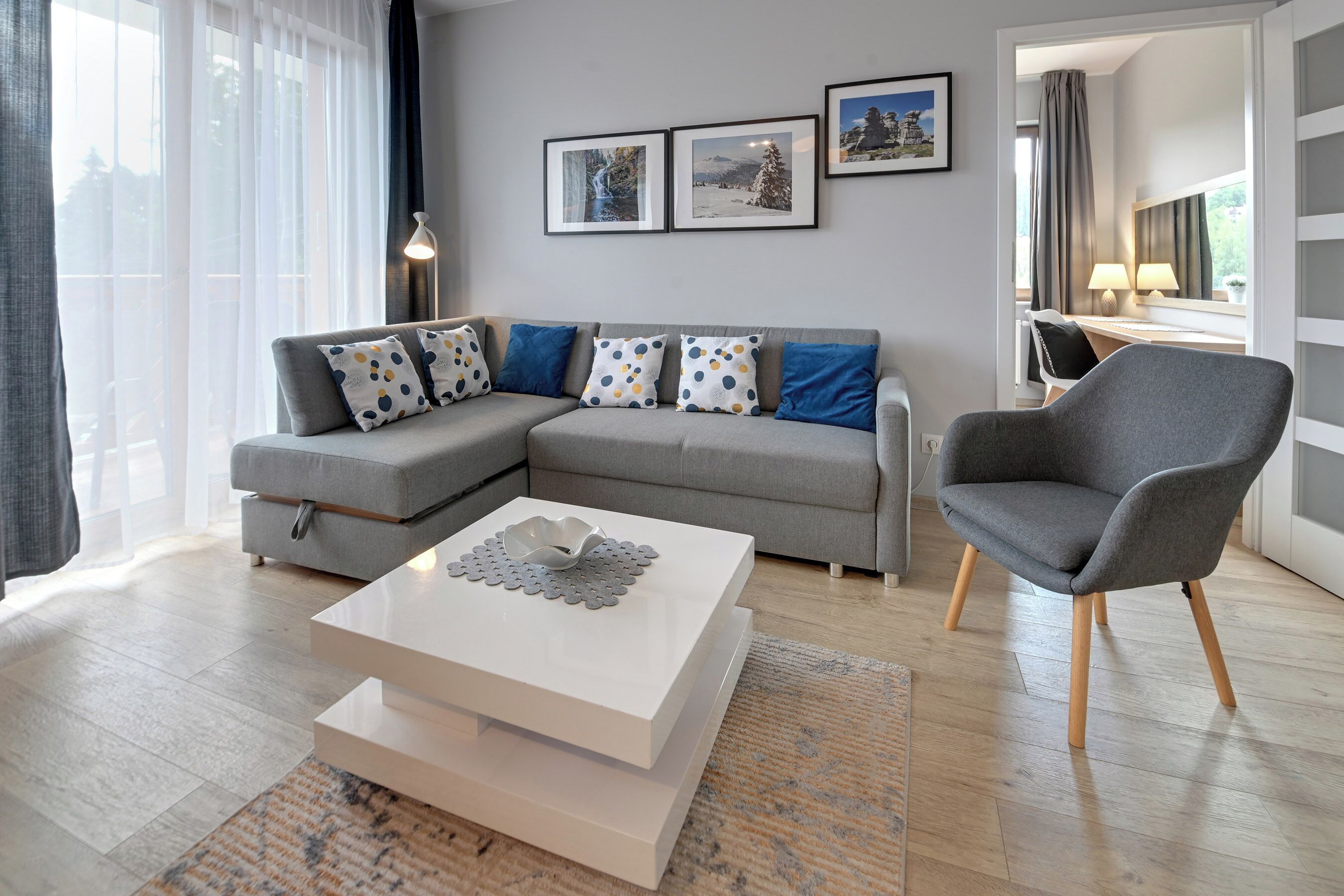 A modern living room with a grey sectional sofa, a white coffee table, and a grey armchair. There are three framed pictures on the wall and a lamp on the side table.