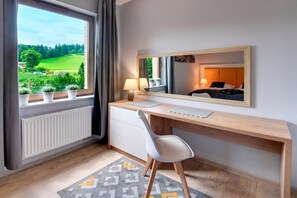 A modern hotel room with a wooden desk, a white chair, a large window with a view of a green landscape, and a mirror reflecting the room. - RentPlanet - Apartamenty Bawaria (Szklarska Poreba)