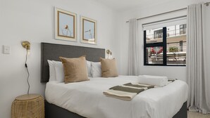 A neatly made bed with a grey headboard, white bedding, and a striped throw blanket. There are two framed pictures on the wall above the bed, a round wicker side table, and a window with curtains. - Azure 32 (Cape Town)