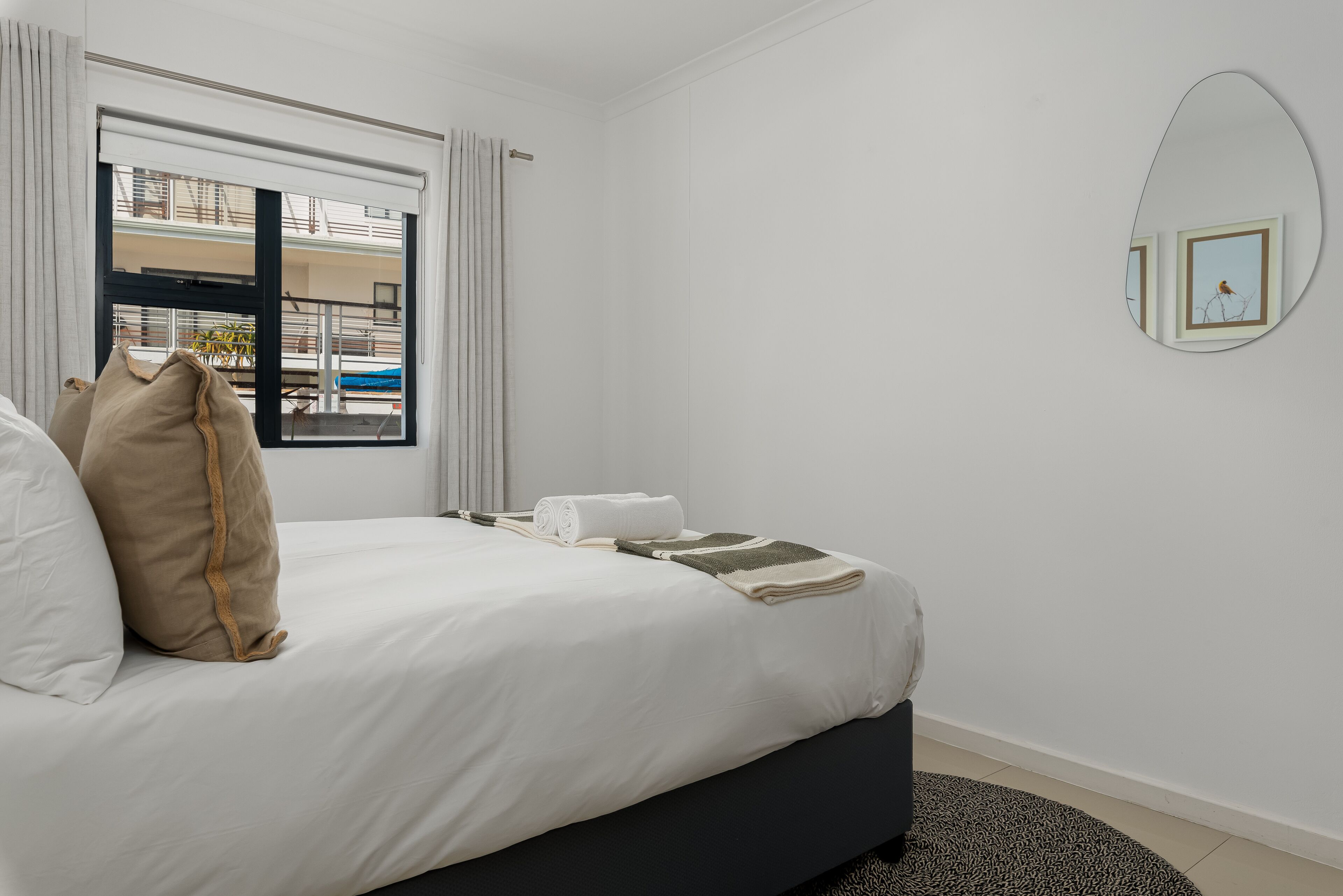 A neatly made bed with white and beige bedding, a dark-colored headboard, and a window with curtains.