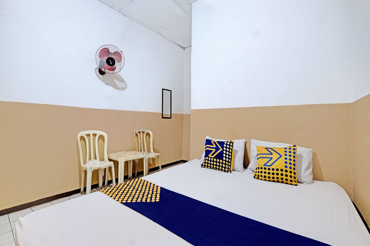 A hotel room with a bed, two chairs, a small table, and a wall-mounted fan.