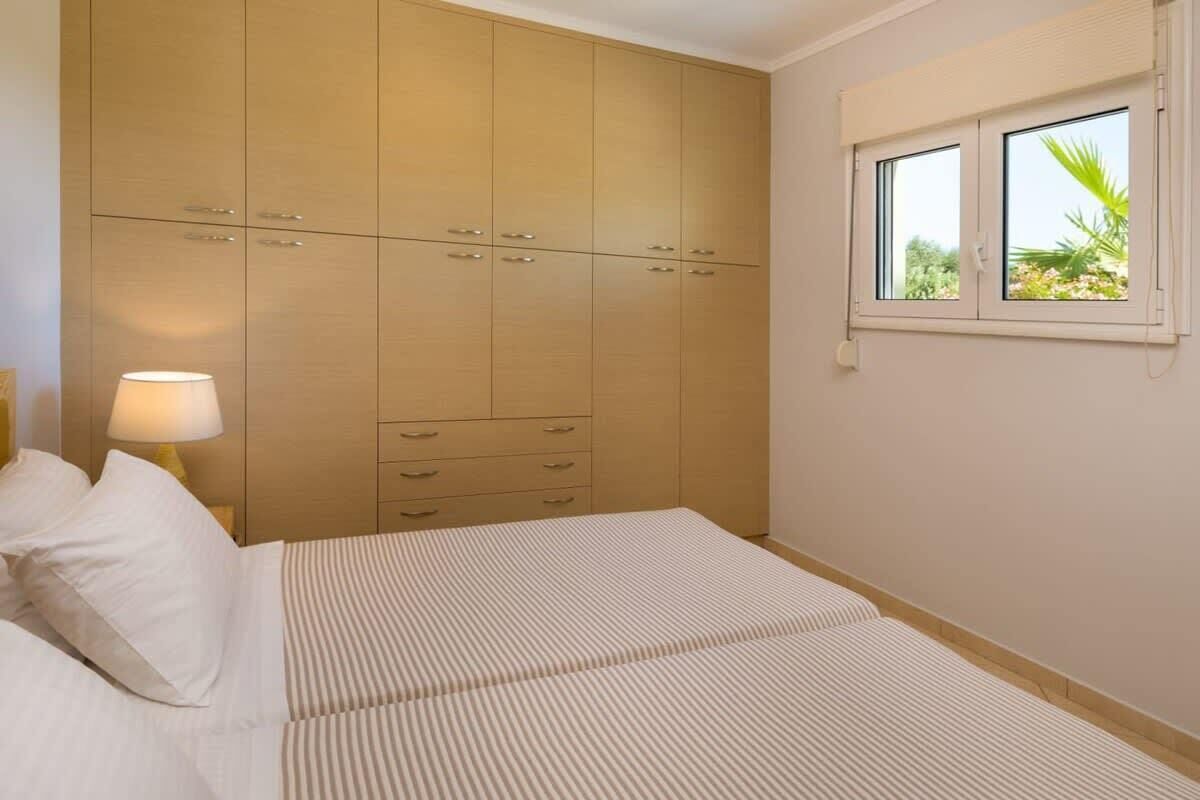 A bedroom with a large wardrobe, a bed with striped bedding, a bedside lamp, and a window with a view of greenery.