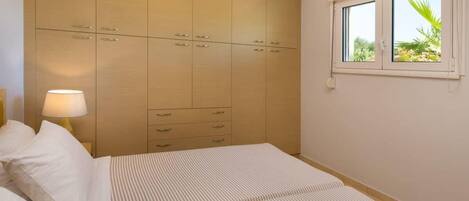 A bedroom with a large wardrobe, a bed with striped bedding, a bedside lamp, and a window with a view of greenery.