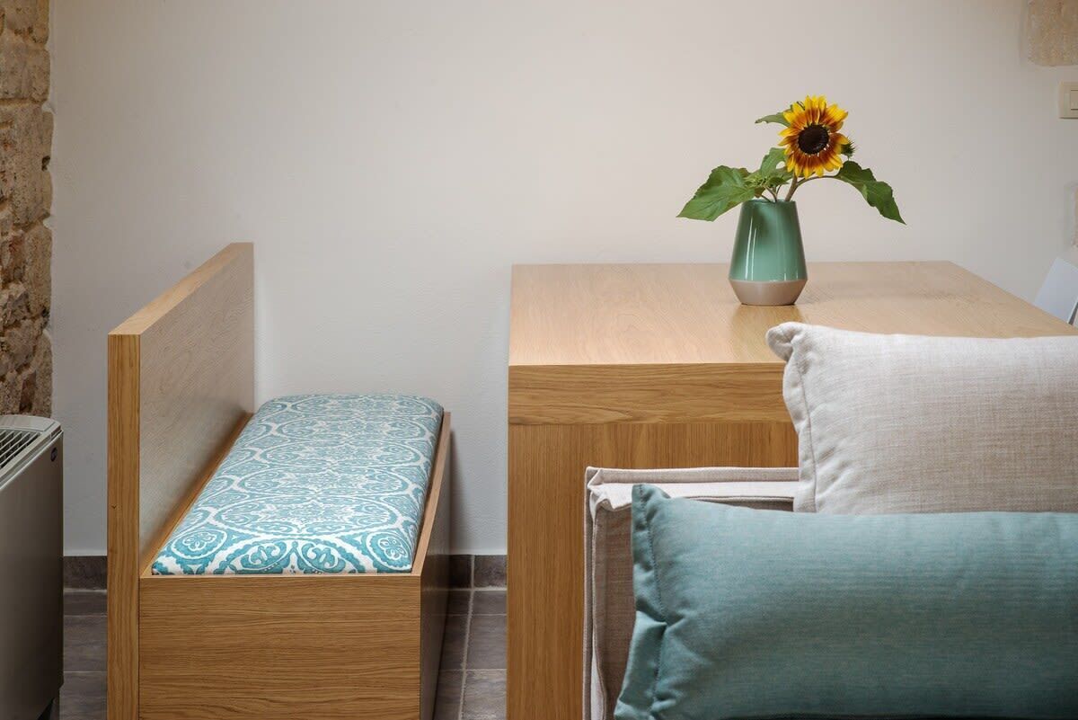 A bedroom with a wooden bed, a bedside table with a vase of sunflowers, and a bench with a patterned cushion.