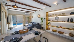 A modern dining area with a wooden table set for four, a sofa in the background, and a shelf with decorative items. - Casa di Grimpili (Kolimvari)
