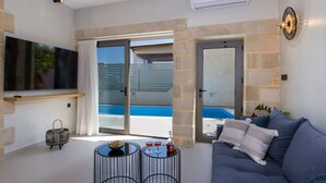 A modern living room with a large TV, a sofa, two small tables, and a view of a pool. - Casa di Grimpili (Kolimvari)