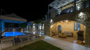 A modern outdoor pool area with lounge chairs, a barbecue grill, and a stone archway leading to a covered seating area. - Casa di Grimpili (Kolimvari)