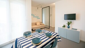 A modern room with a dining table set for four, a sofa, a TV mounted on the wall, and a large window with blinds.