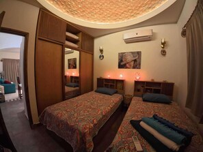A room with two beds, a wardrobe, a mirror, an air conditioner, and a painting on the wall.
