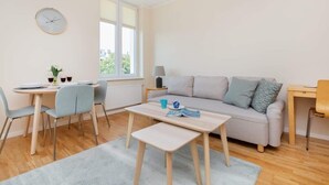 En moderne stue med sofa, spisebord og stole. Et ur hænger på væggen, og et vindue giver naturligt lys.