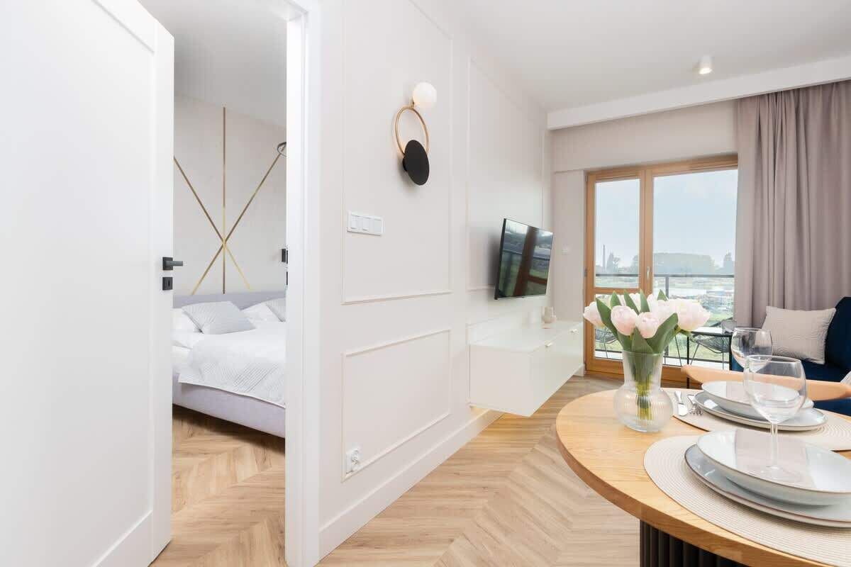 Quarto de hotel moderno com cama, área de refeições com mesa posta para dois, televisão fixada na parede e uma grande janela com vista.