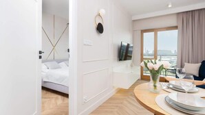 A modern hotel room with a bed, a dining area with a table set for two, a TV mounted on the wall, and a large window with a view. - Elegant Apartment | Balcony with View of the Motława River, Gymnasium (Gdansk)