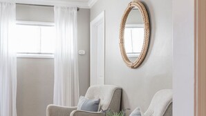 Una habitación con dos sillones, un espejo tejido y una ventana con cortinas blancas.