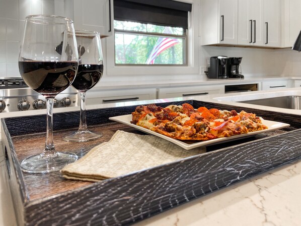 A kitchen counter with a plate of food, two glasses of red wine, and a window with a view of trees.