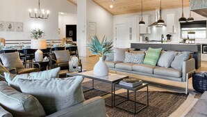 A modern living room with a sofa, coffee table, and dining area. There is a kitchen in the background with white cabinets and a black countertop. - Luxury at Lake Taneycomo, Hot Tub, FirePit, Views! (Hollister)