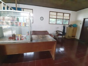 Ein Zimmer mit einem großen Kühlschrank, der mit Getränken gefüllt ist, einem Holztisch, einem Korbstuhl, einem kleinen Tisch mit Stuhl und einem Fenster mit Jalousien.