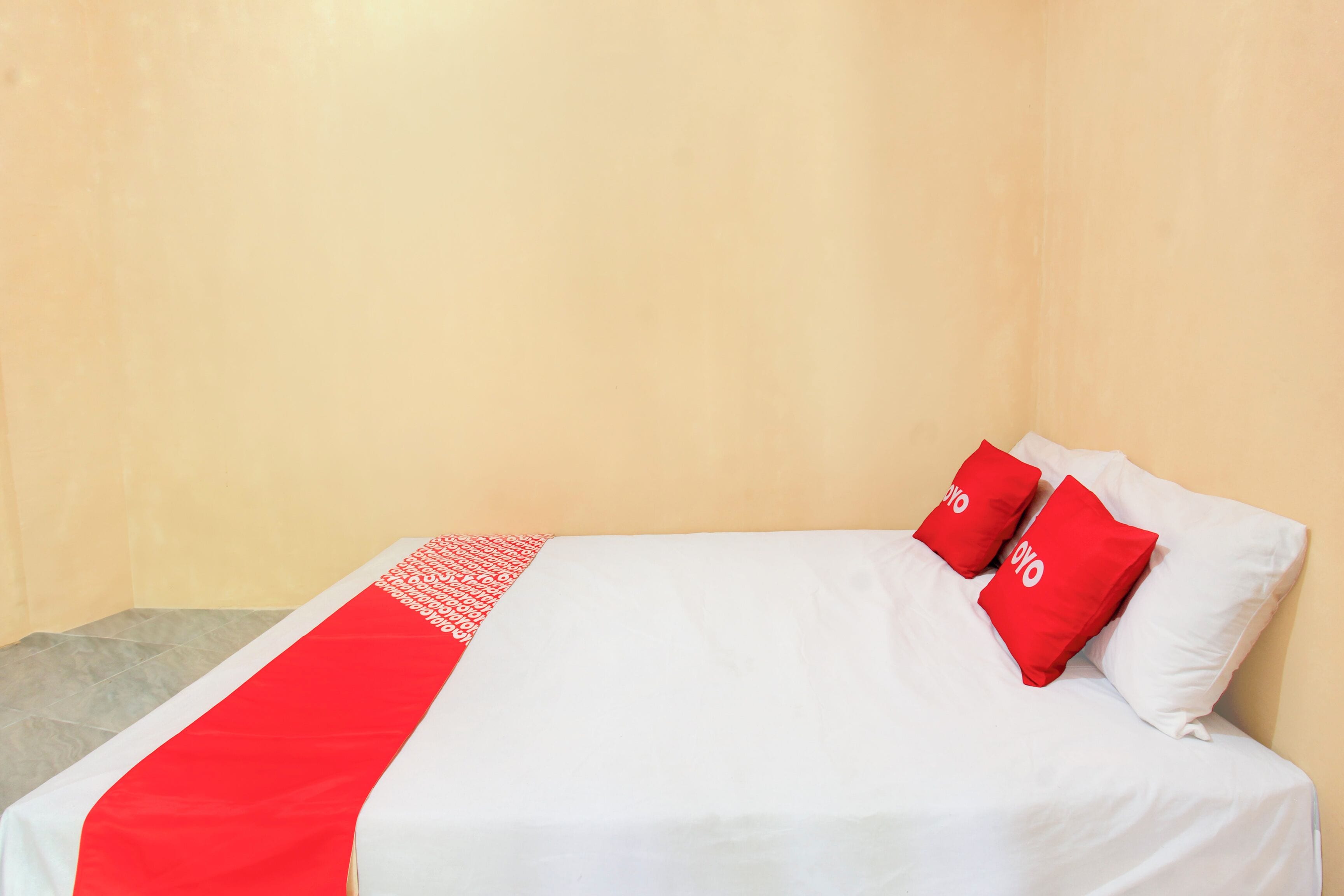 A single bed with white bedding and two red pillows, a grey rug, and a plain wall.
