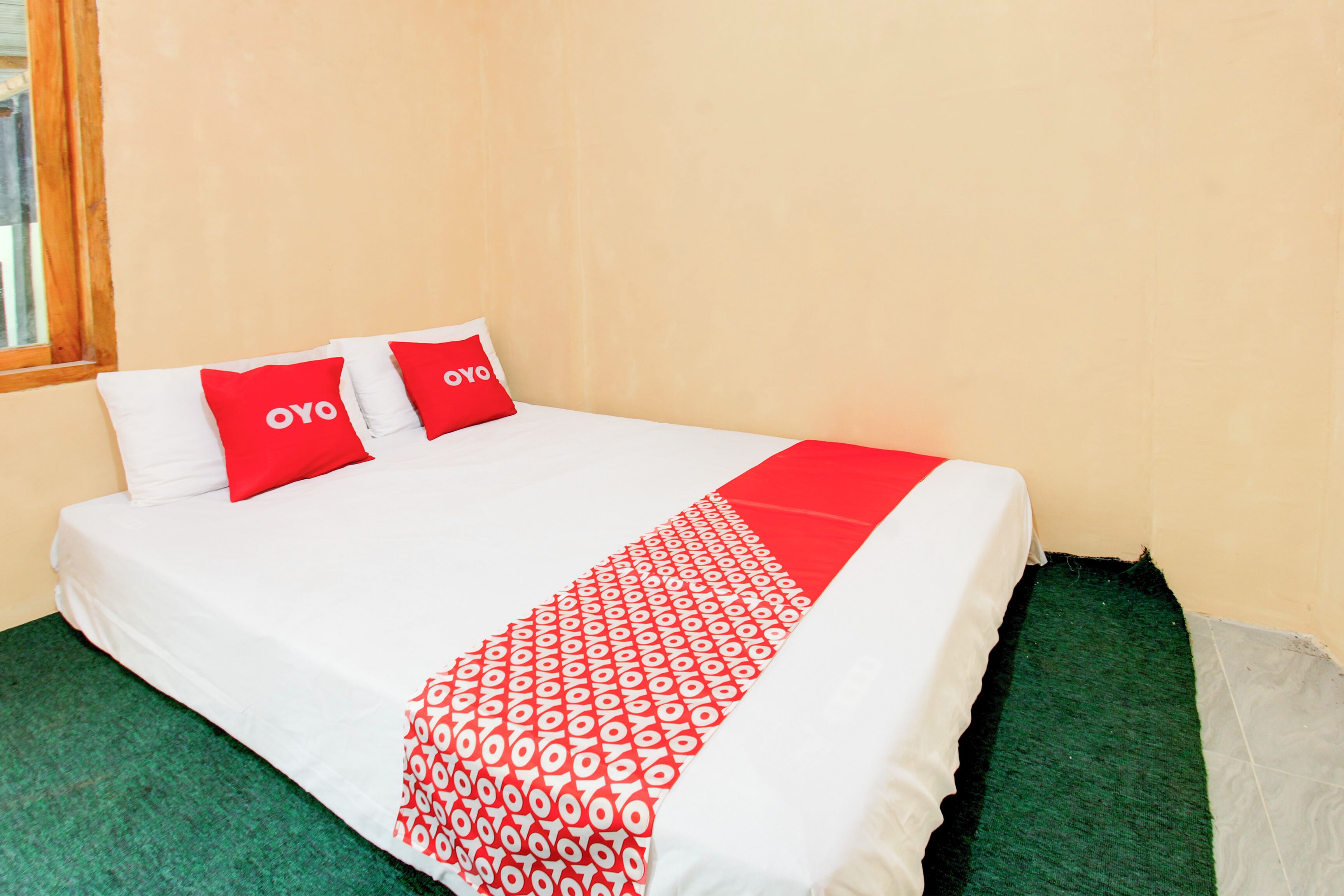 Una cama con ropa blanca y almohadas rojas, un plaid rojo y blanco, y alfombra verde.