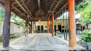 An outdoor covered seating area with wooden benches and a tiled floor, surrounded by trees and a building with a yellow facade. - Hotel O Kost Gayus (Mataram)