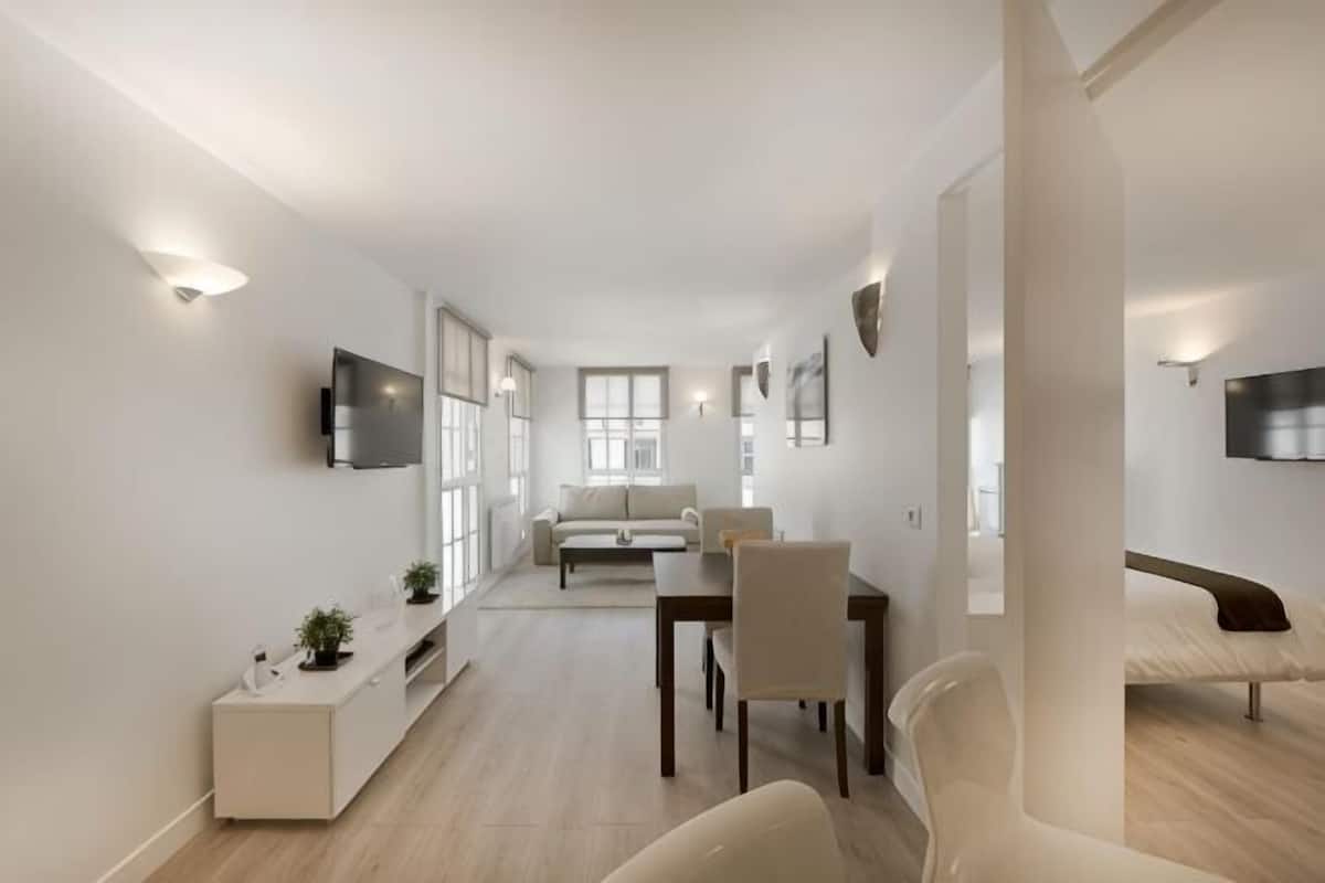 Una sala de estar moderna con comedor, televisor montado en la pared y una cama al fondo.