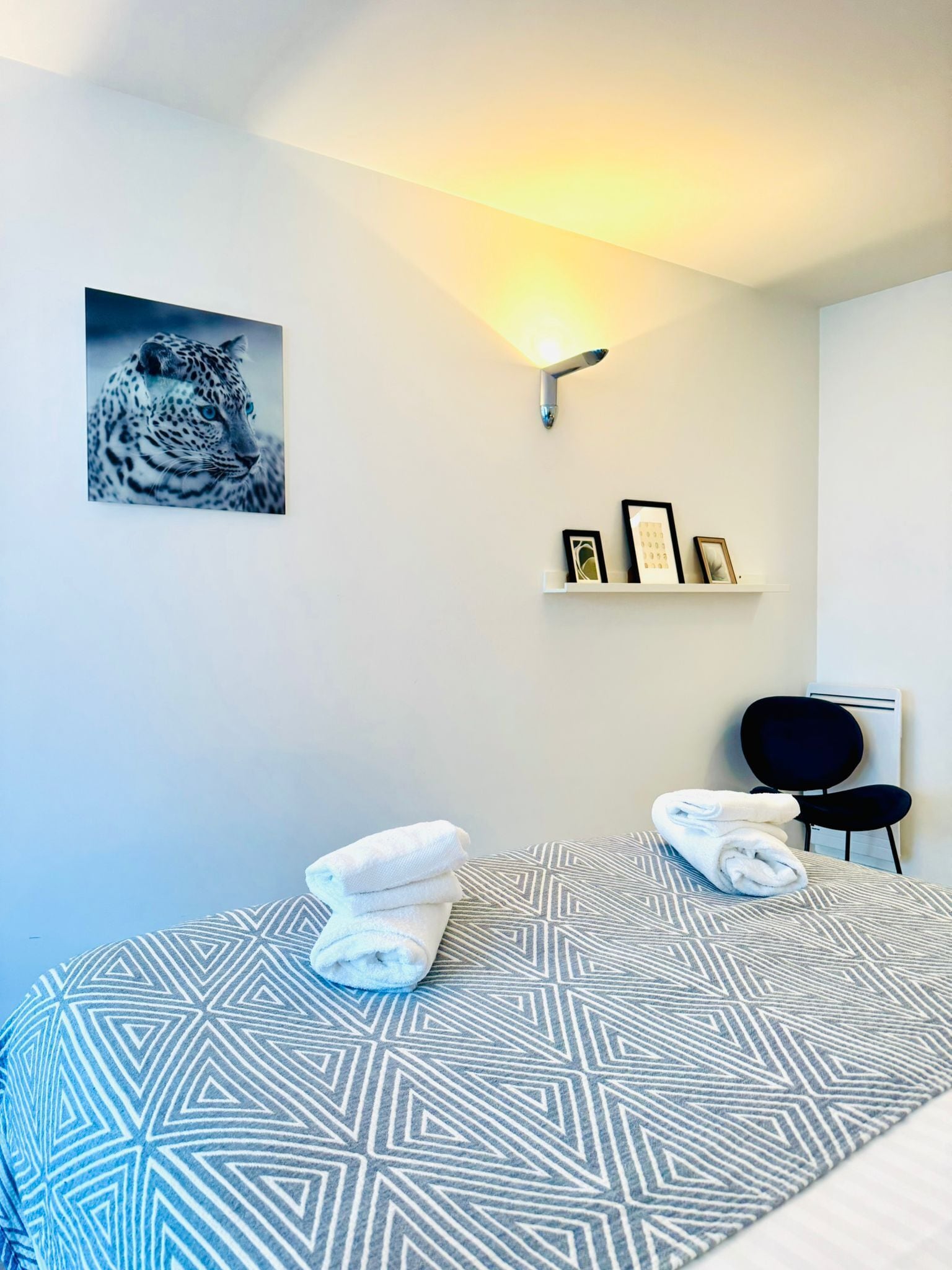 Quarto de hotel com uma cama, um quadro de leopardo na parede, uma prateleira com fotos emolduradas e uma cadeira.