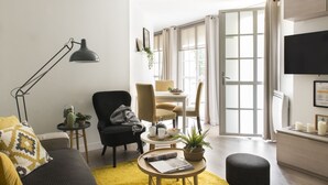 A modern living room with a black armchair, a yellow sofa, a round coffee table, and a floor lamp. There is a dining area with a table and chairs, and a TV mounted on the wall. - Le Logis Versaillais (Versailles)